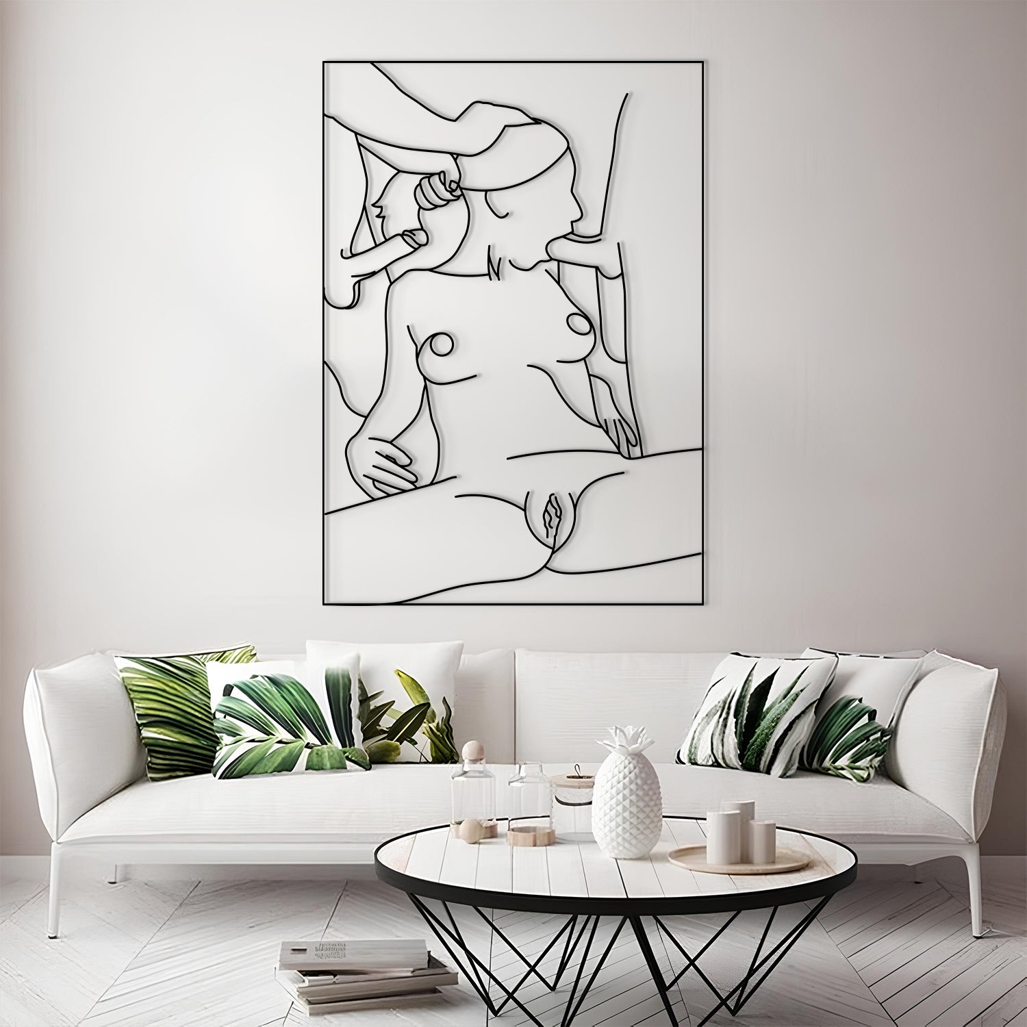 A framed minimalist black line art print of a nude female figure is displayed on a light gray wall above a white sofa with green leaf-patterned pillows in a modern living room setting.