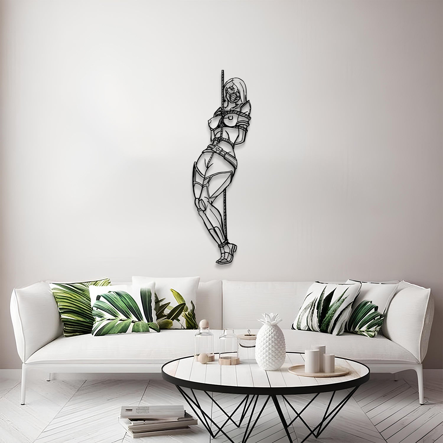 A black metal wall art piece depicting a stylized, geometric female figure hanging from a pole, displayed on a light gray wall above a modern white sofa with green leaf-patterned pillows.