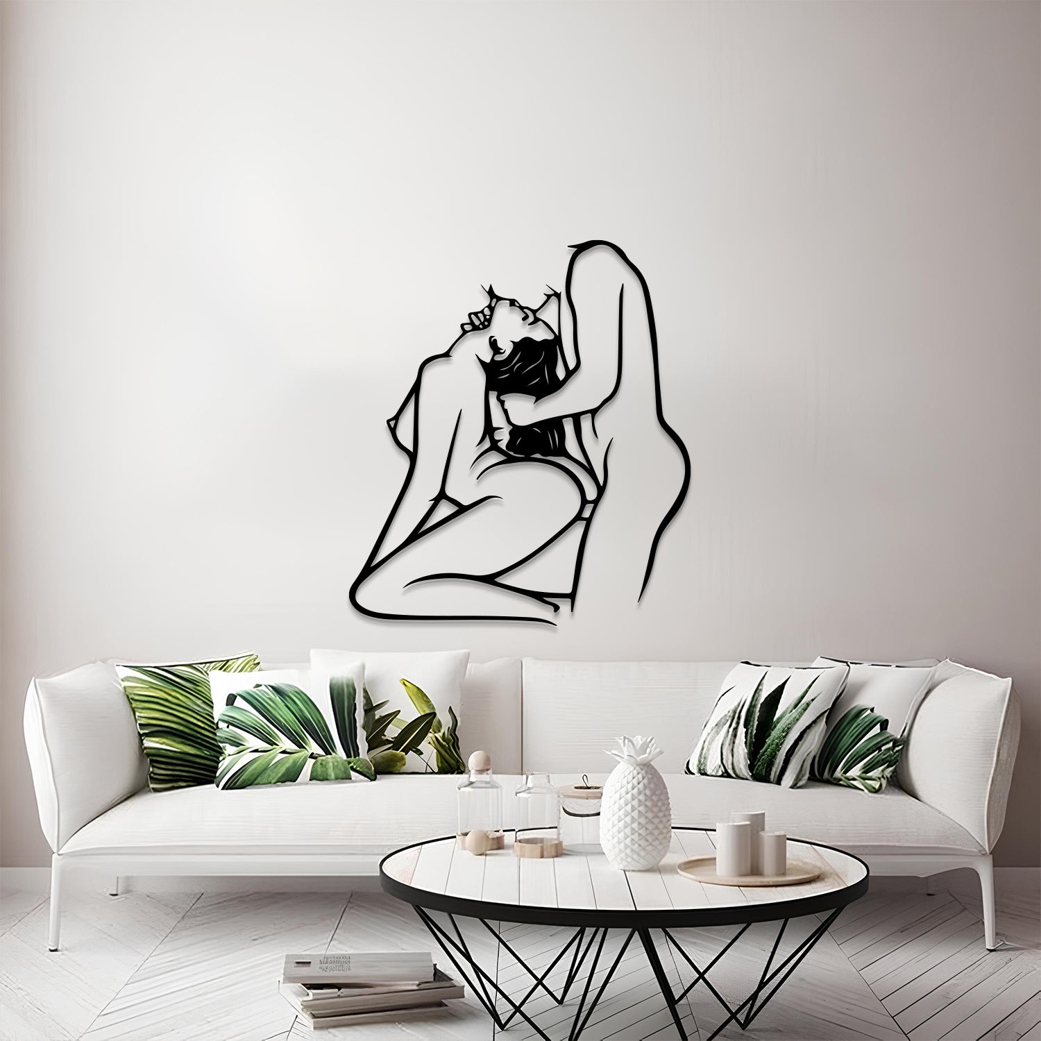 A black metal wall art piece depicting a minimalist line drawing of a kissing couple, mounted on a light gray wall above a modern white sofa with green leaf-patterned pillows.