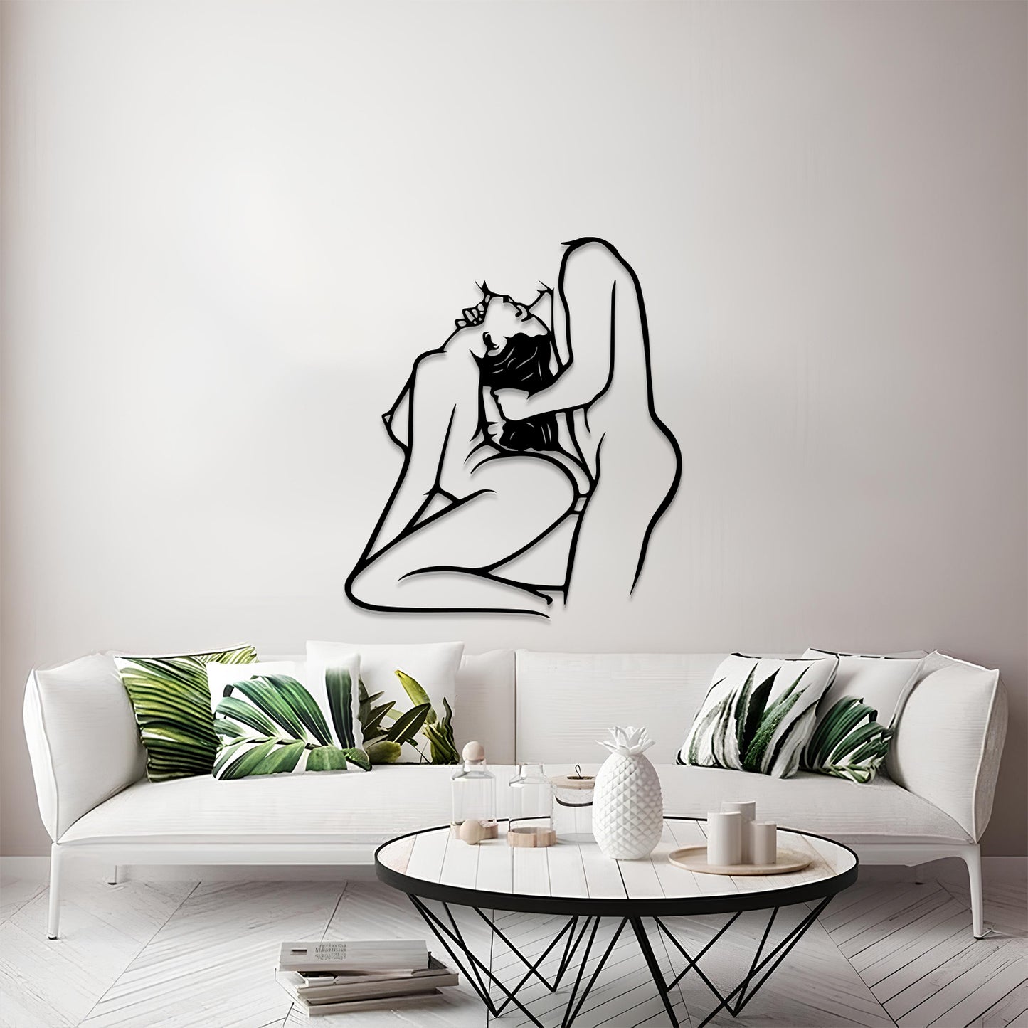 A black metal wall art piece depicting a minimalist line drawing of a kissing couple, mounted on a light gray wall above a modern white sofa with green leaf-patterned pillows.