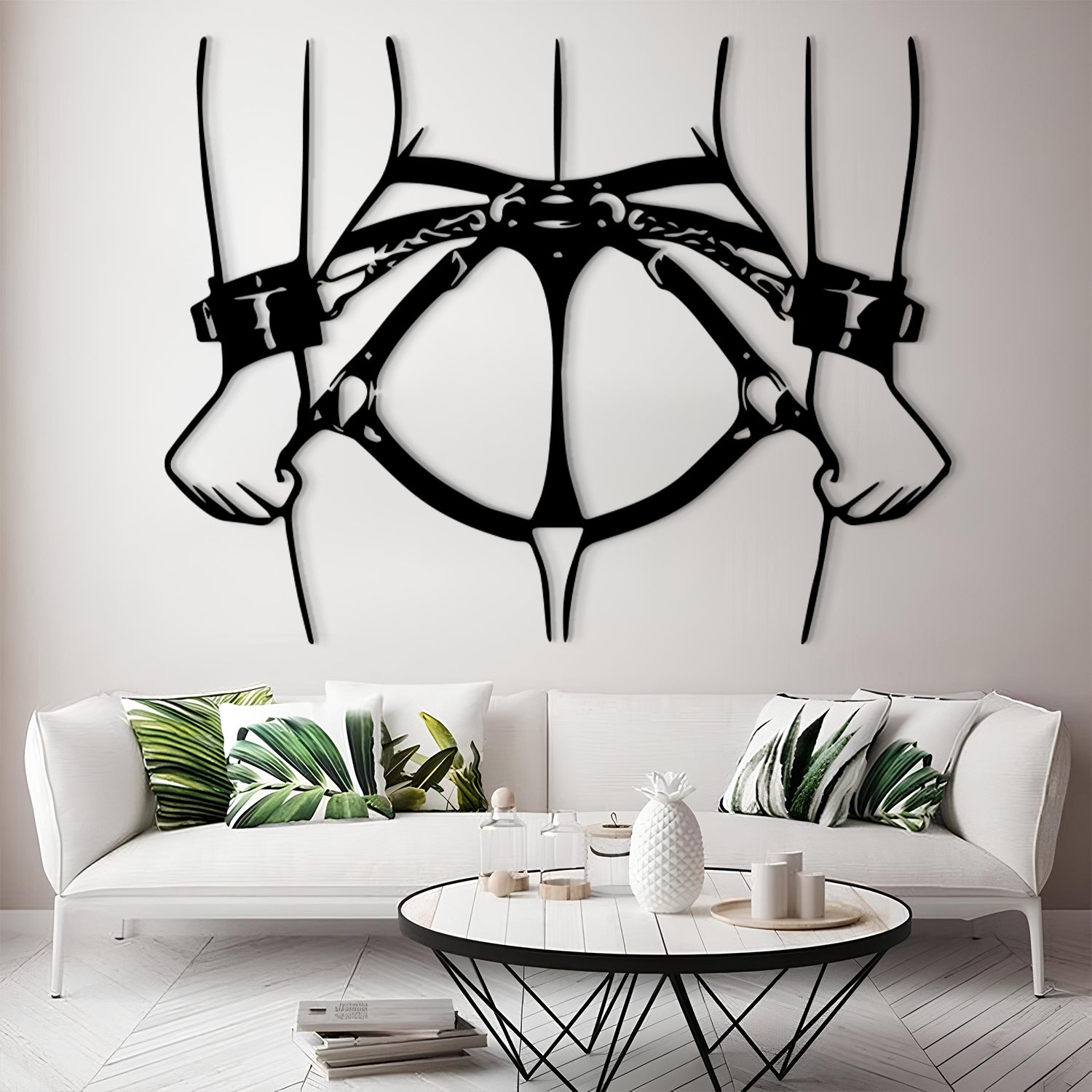 A black metal wall art piece depicting a harness and two hands, displayed on a light gray wall above a modern white sofa with green leaf-patterned pillows.