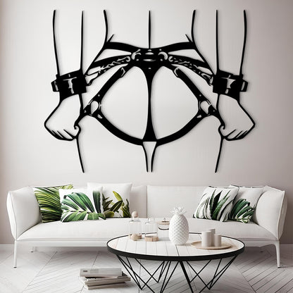 A black metal wall art piece depicting a harness and two hands, displayed on a light gray wall above a modern white sofa with green leaf-patterned pillows.