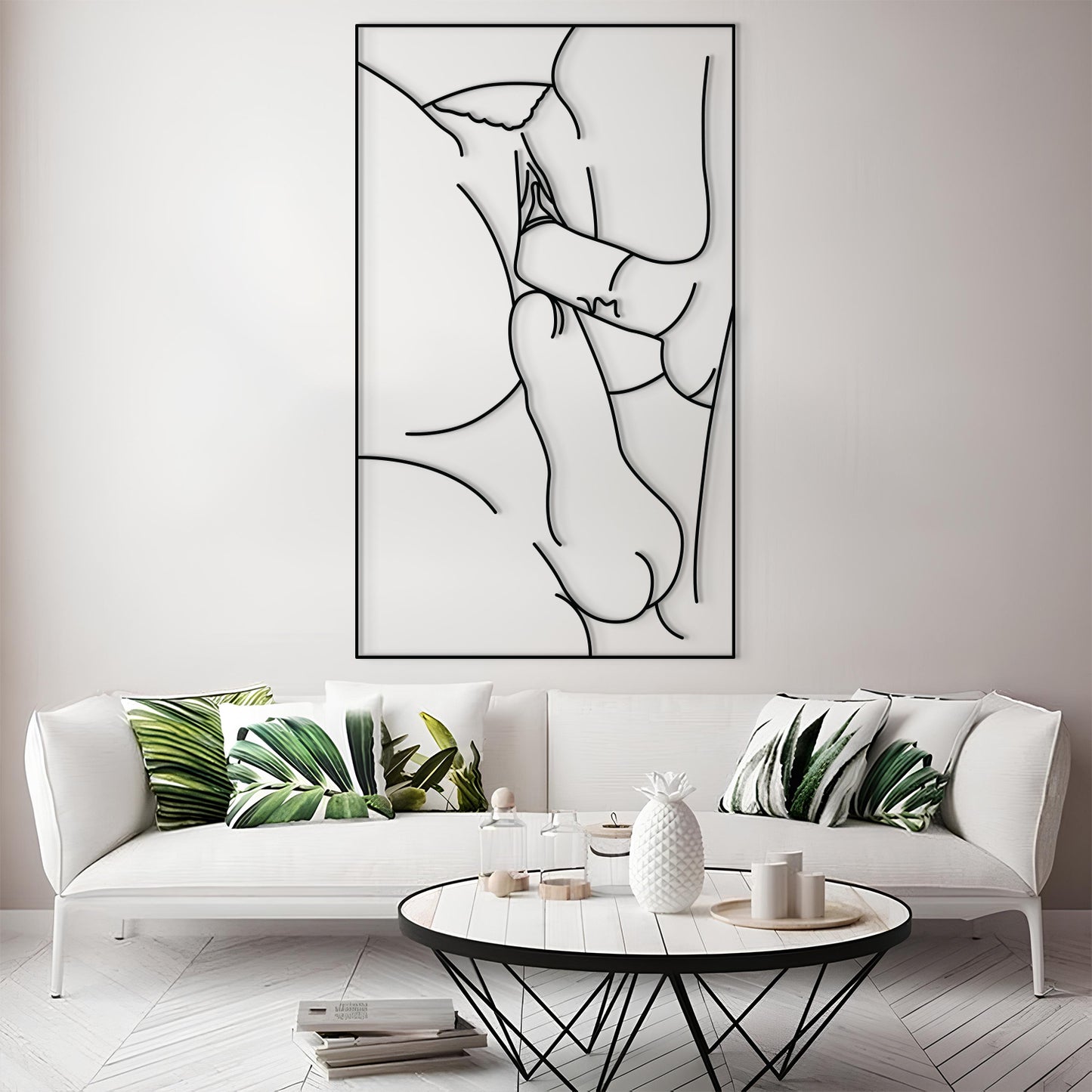 A framed minimalist black line art print of a couple embracing, displayed on a light gray wall above a modern white sofa with green leaf-patterned pillows in a bright living room.
