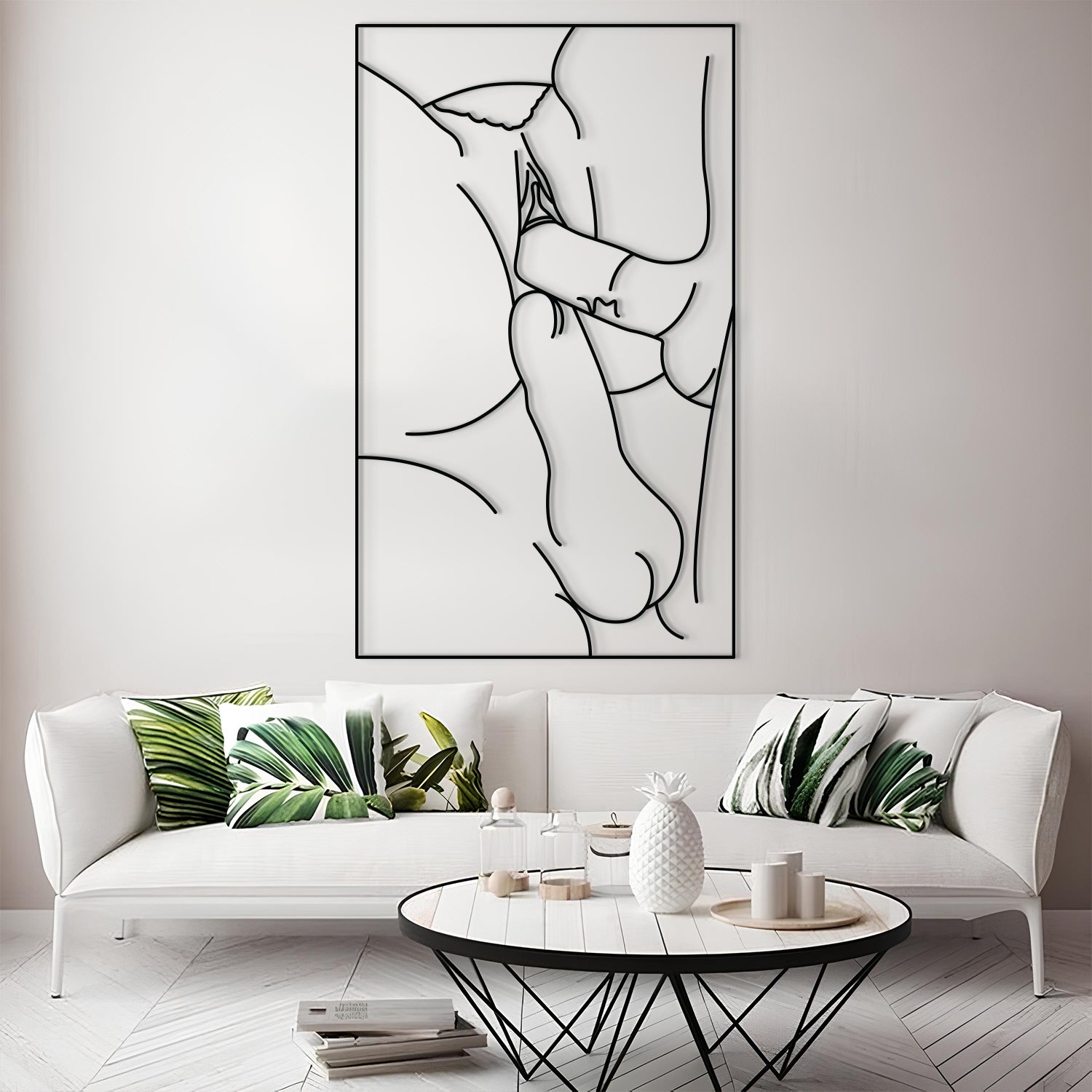 A framed minimalist black line art print of a couple embracing, displayed on a light gray wall above a modern white sofa with green leaf-patterned pillows in a bright living room.