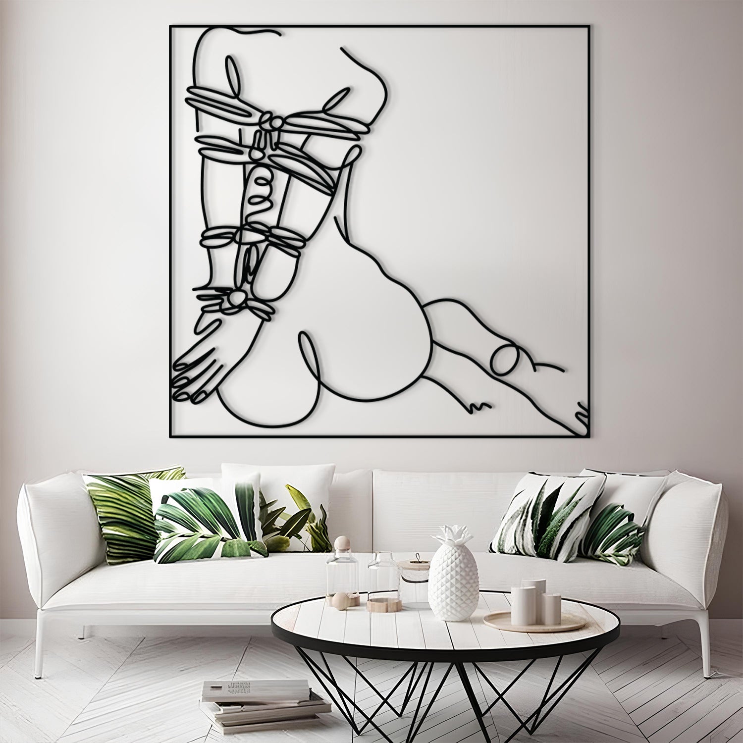 A large black and white minimalist line art wall hanging depicting a censored torso, displayed above a white sofa with green leaf-patterned pillows in a modern living room setting.