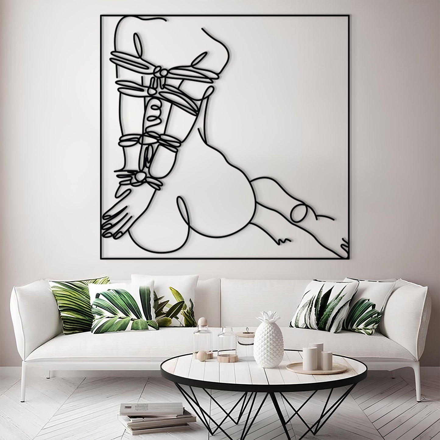 A large black and white minimalist line art wall hanging depicting a censored torso, displayed above a white sofa with green leaf-patterned pillows in a modern living room setting.