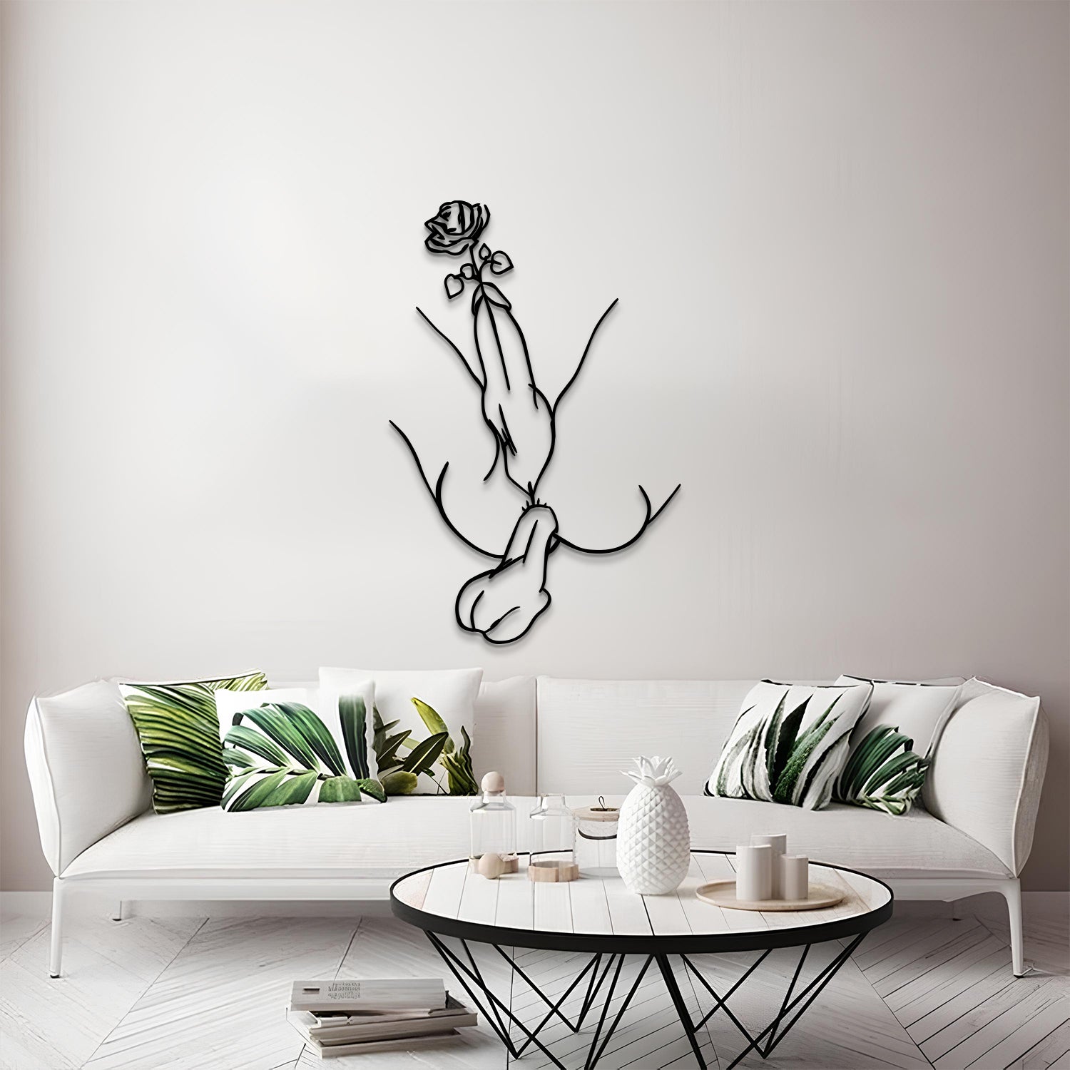 A modern living room scene featuring a black metal line art wall sculpture of a hand holding a rose, mounted on a light gray wall above a white sofa with green leaf-patterned pillows.