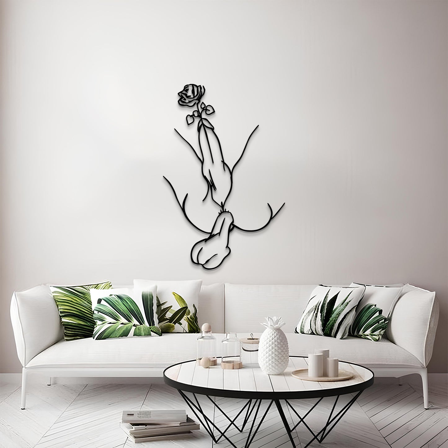 A modern living room scene featuring a black metal line art wall sculpture of a hand holding a rose, mounted on a light gray wall above a white sofa with green leaf-patterned pillows.