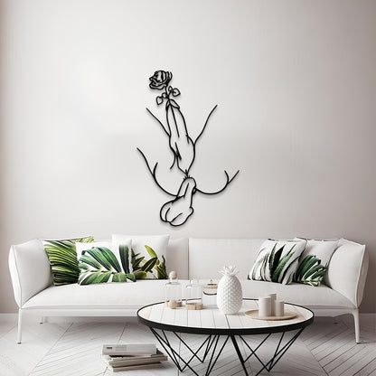 A modern living room scene featuring a black metal line art wall sculpture of a hand holding a rose, mounted on a light gray wall above a white sofa with green leaf-patterned pillows.