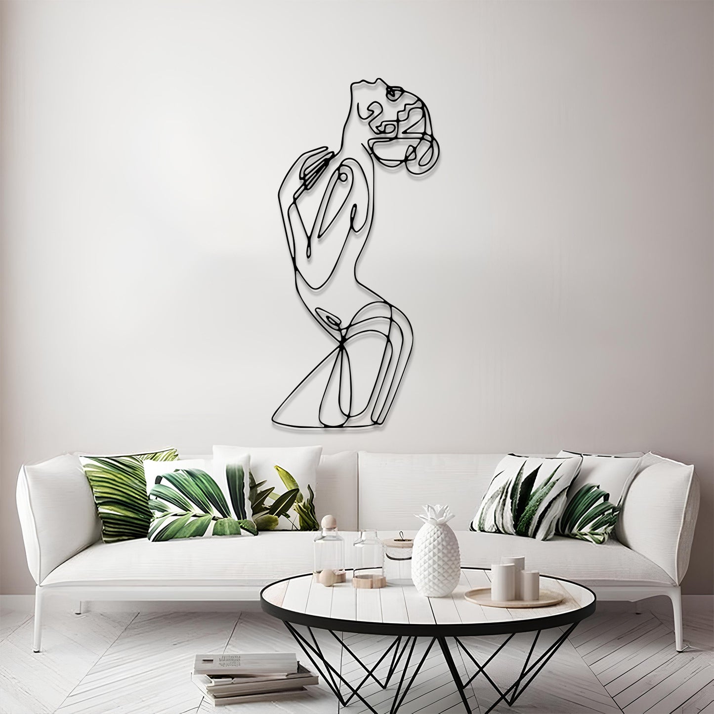 A modern minimalist living room featuring a black wire art piece of a stylized female figure on the wall above a white sofa with green leaf-patterned pillows.