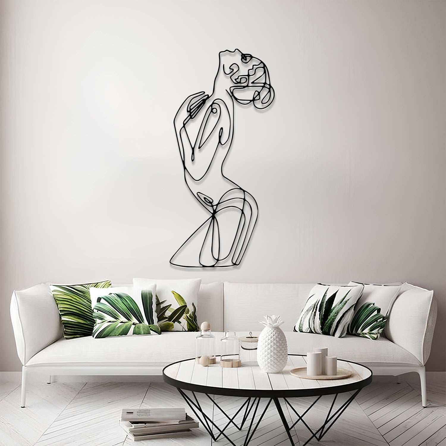 A modern minimalist living room featuring a black wire art piece of a stylized female figure on the wall above a white sofa with green leaf-patterned pillows.