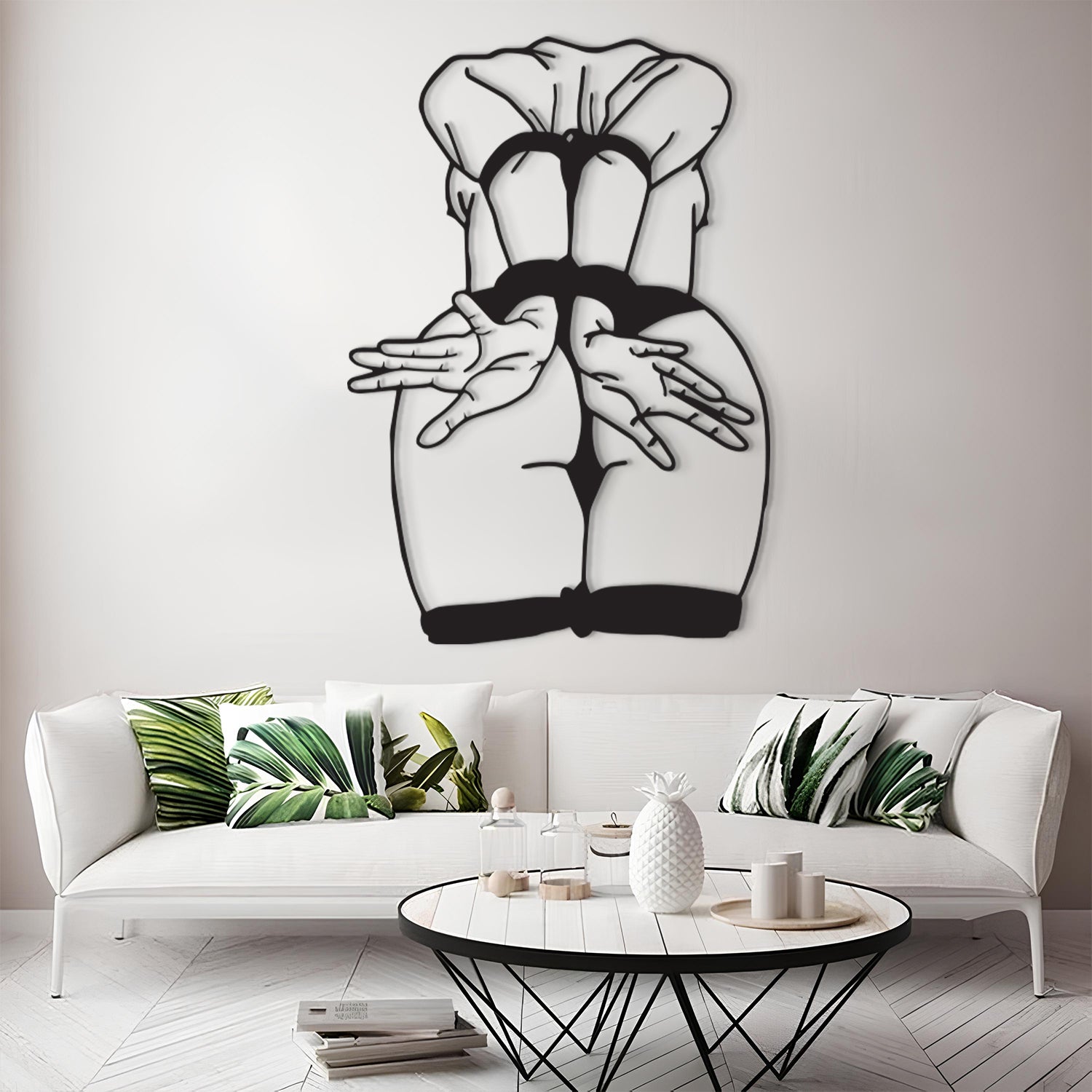 A black metal wall art piece depicting a stylized rear view of a woman in a chef's hat and black dress, displayed on a light gray wall above a modern white sofa with green leaf-patterned pillows.