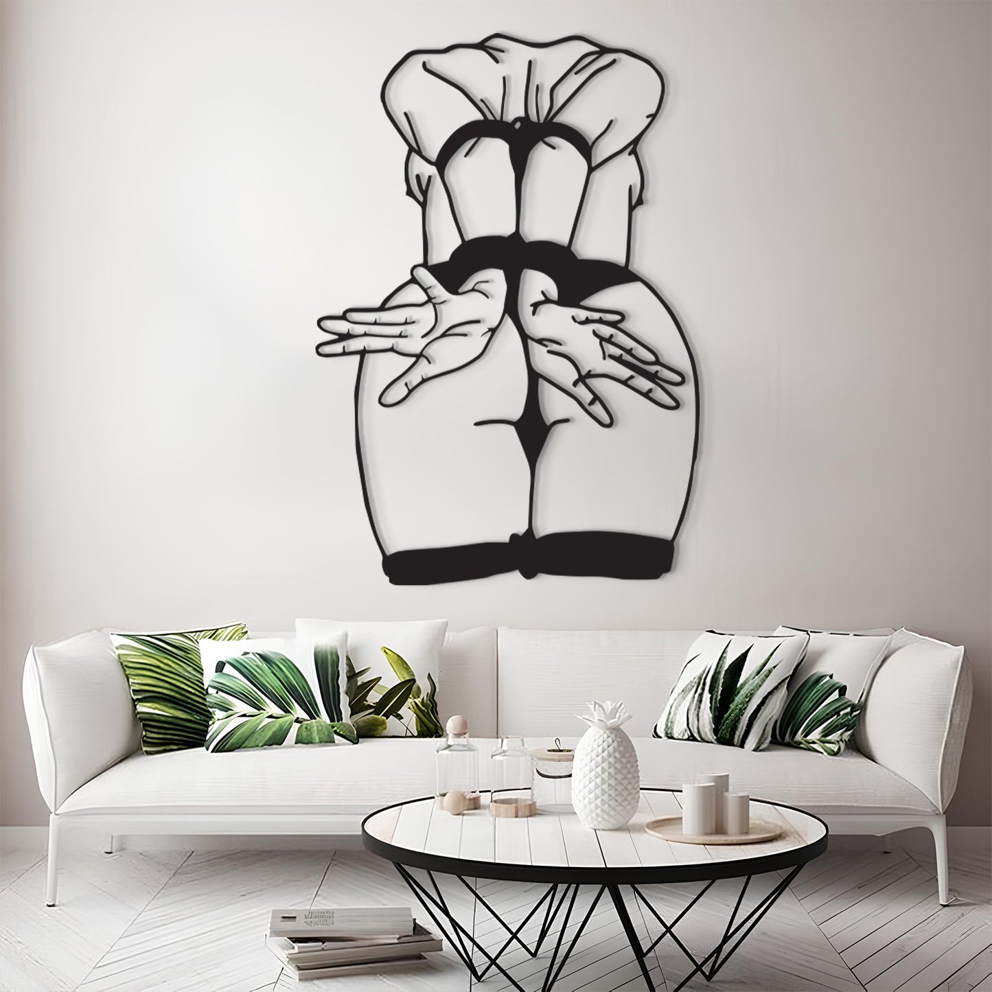 A black metal wall art piece depicting a stylized rear view of a woman in a chef's hat and black dress, displayed on a light gray wall above a modern white sofa with green leaf-patterned pillows.
