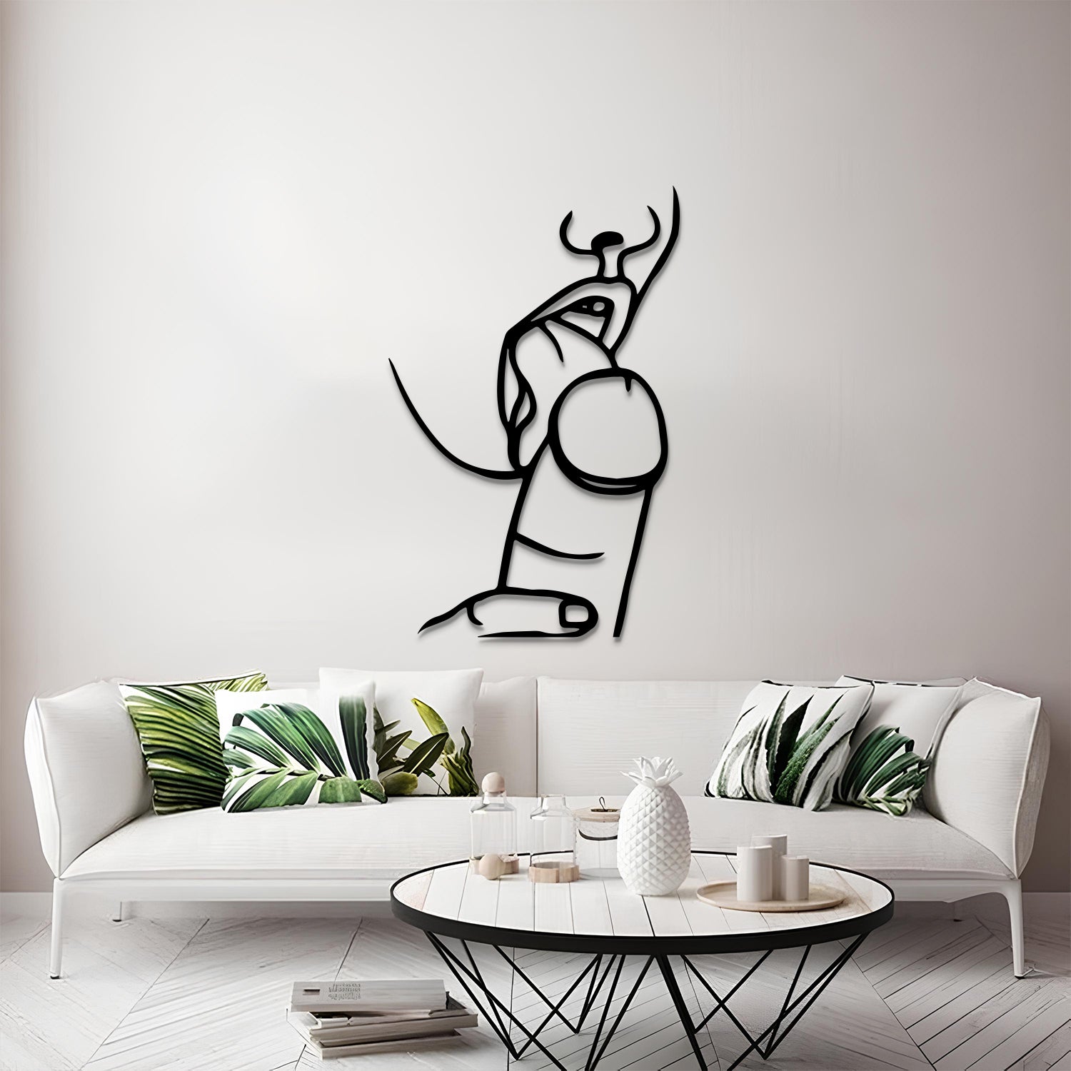 A minimalist black metal wall art piece depicting an abstract bull, displayed on a light gray wall above a modern white sofa with green leaf-patterned pillows.