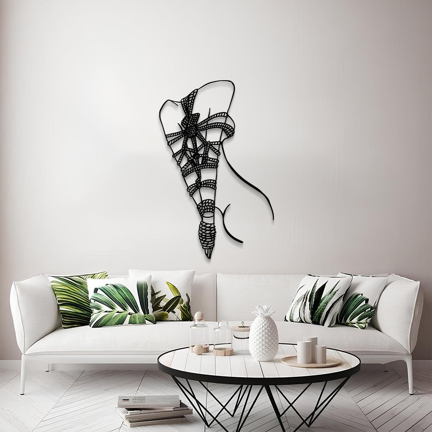 A modern living room scene featuring a black metal wire wall art piece shaped like a stylized ribbon or bow, mounted on a light gray wall above a white sofa with green leaf-patterned pillows.