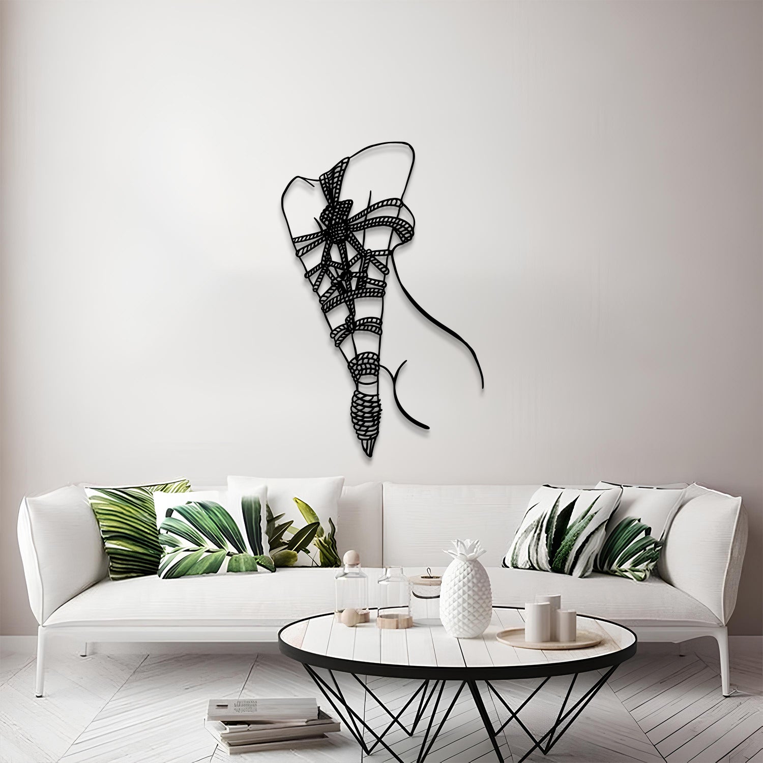 A modern living room scene featuring a black metal wire wall art piece shaped like a stylized ribbon or bow, mounted on a light gray wall above a white sofa with green leaf-patterned pillows.