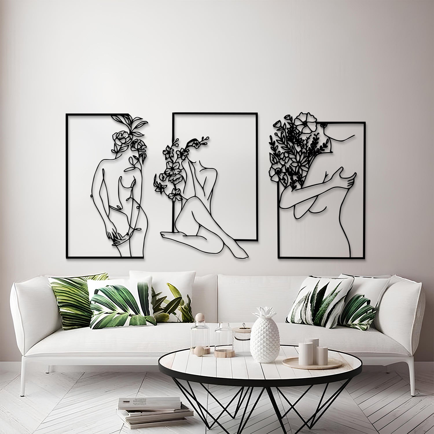 Three framed black metal wall art prints depicting minimalist line-art female figures with flowers, displayed above a white sofa in a modern living room setting.