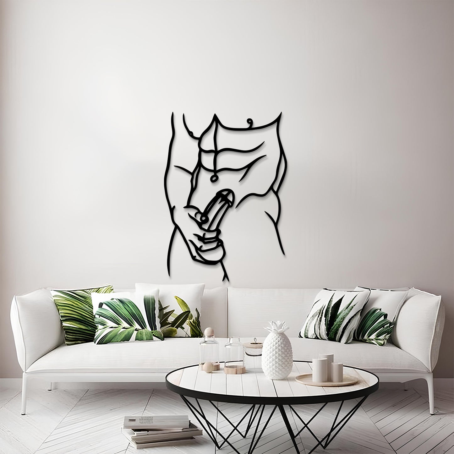 A modern living room scene featuring a black metal wall art piece of a muscular torso with a peace sign, mounted on a light gray wall above a white sofa with green leaf-patterned pillows.