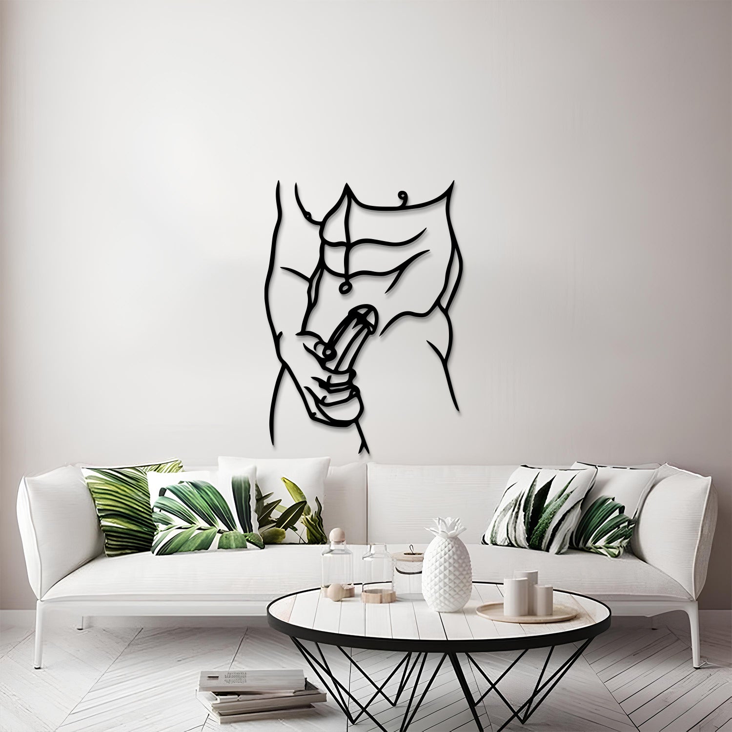 A modern living room scene featuring a black metal wall art piece of a muscular torso with a peace sign, mounted on a light gray wall above a white sofa with green leaf-patterned pillows.