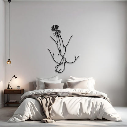 A minimalist bedroom scene with a bed dressed in white and beige linens, a small wooden nightstand with a lamp, and a black line-art wall sculpture of hands holding a rose mounted on the wall above the bed.