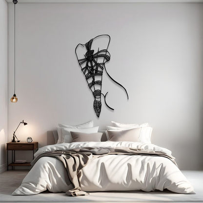 A modern bedroom scene with a bed dressed in white and beige bedding, a small wooden nightstand with a lamp, and a black wire wall art piece shaped like a stylized ribbon bow on the wall above the bed.